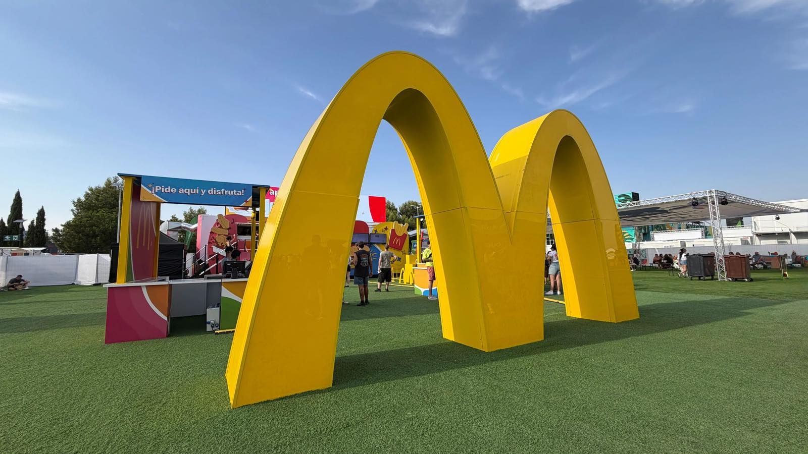 Arcos dorados gigantes de McDonald's instalados sobre césped artificial en la zona de un festival, con visitantes y mostradores de pedido al fondo