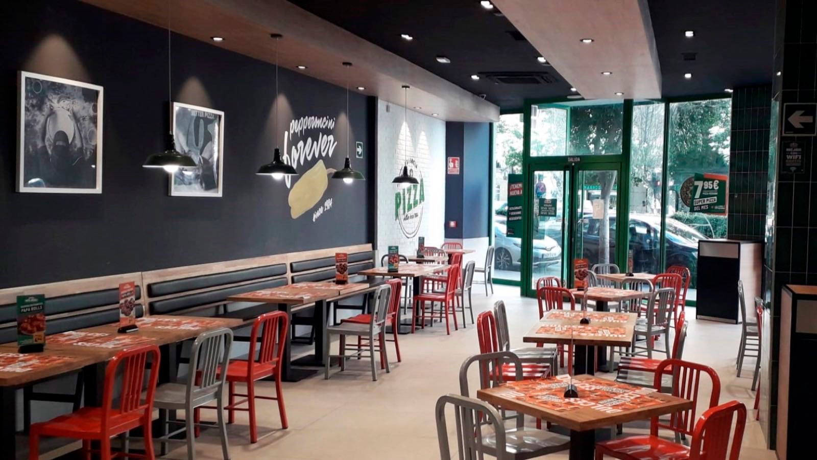 Comedor de una pizzería Papa Johns en España, con mesas de madera, sillas rojas y grandes ventanales a la calle