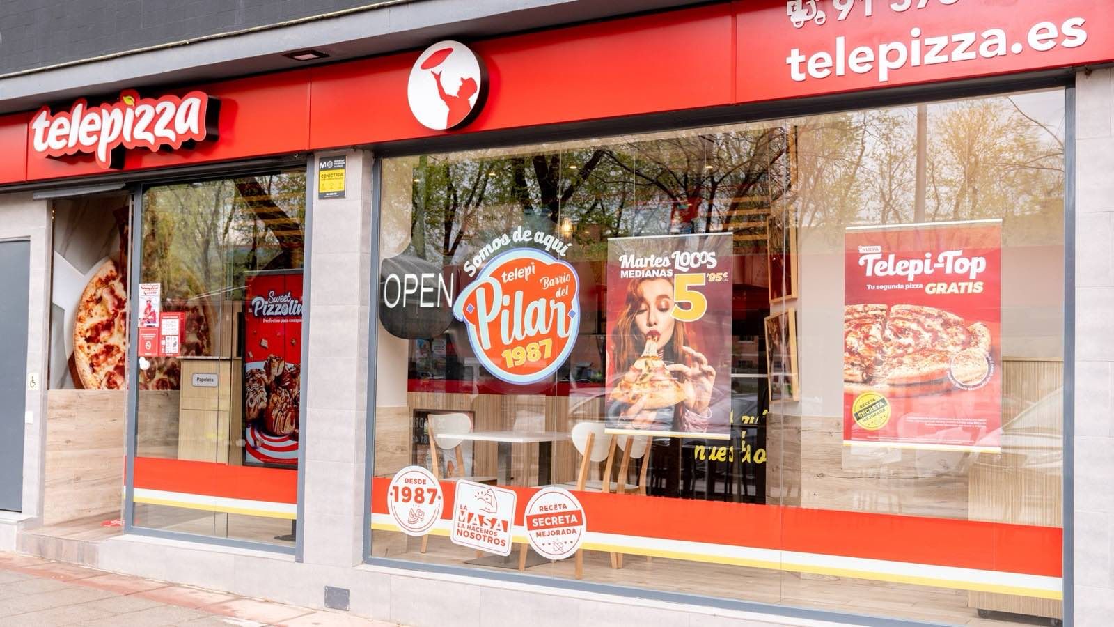 Fachada de una tienda Telepizza con el rótulo rojo de la marca y carteles de promociones en el escaparate
