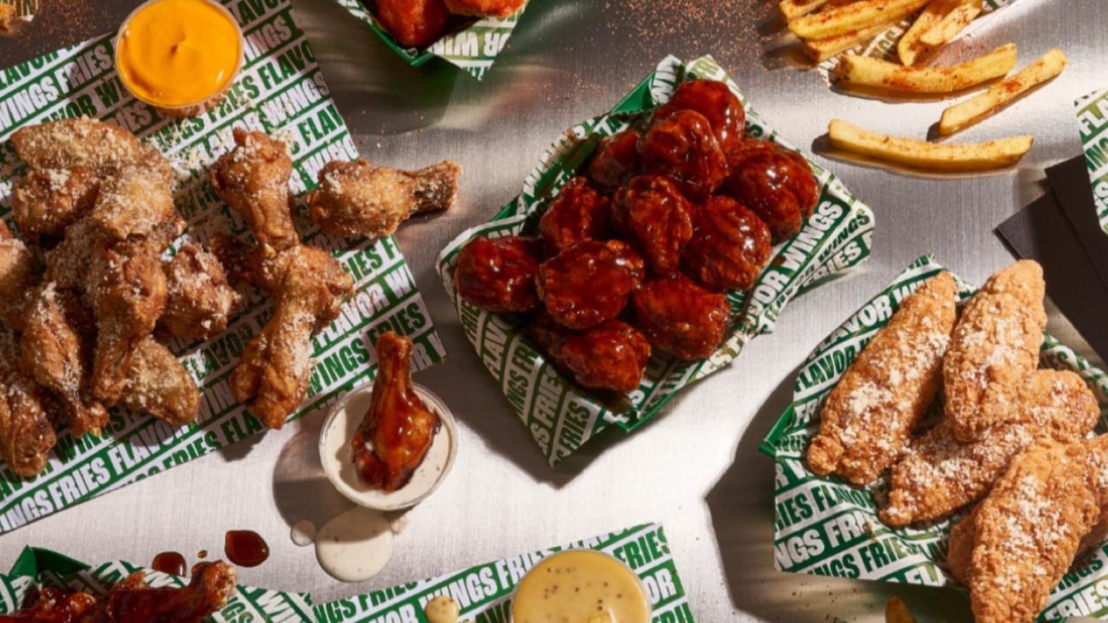 Cestas de alitas, tenders y patatas fritas de Wingstop sobre papel de marca en una mesa metálica